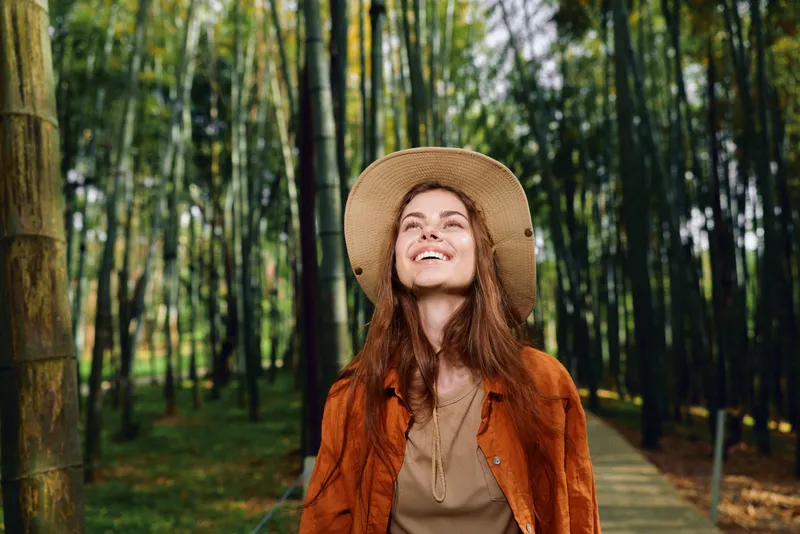 Woman smile hat bamboo forest pathway nature portrait of a young woman walking outdoors on a wooden boardwalk, relaxed and joyful, looking up at sunlight through green foliage.