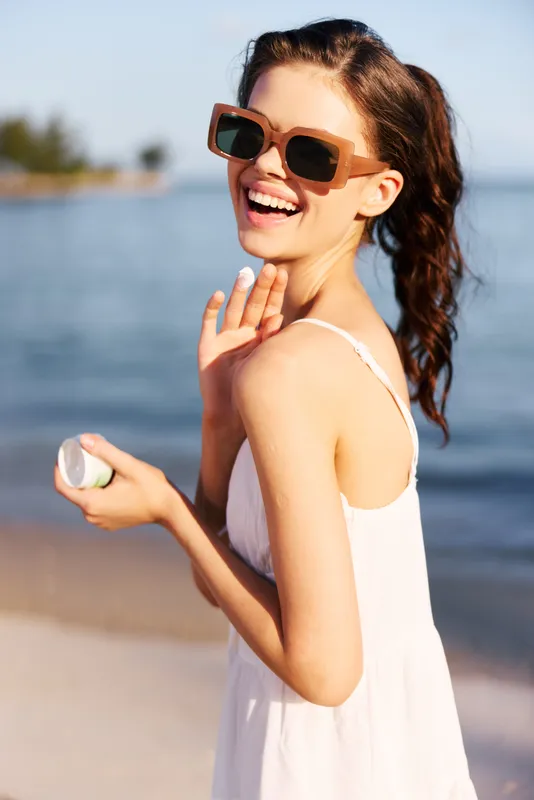 Bright portrait of a smiling woman on the beach wearing sunglasses and a white sundress, holding a shell, with sunlit water and distant shore in the background.