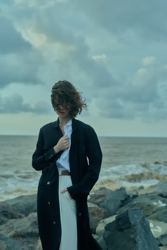 Woman in a long dark coat stands on a rocky shoreline as wind blows through her hair, creating a moody coastal fashion scene.