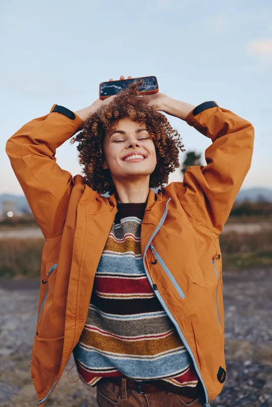 Woman lifestyle outdoors, smiling selfie moment, holding smartphone with curly hair, wearing orange jacket and striped sweater, standing on road background, cheerful travel mood today