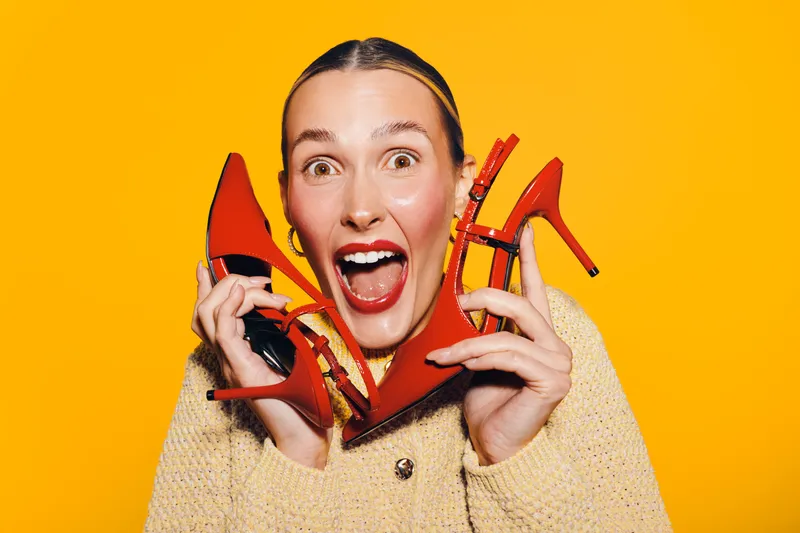 Excited woman with red lipstick holds red high heels near face on bright yellow background. Concept of fashion, style, shoes, and joyful emotions in studio lighting.