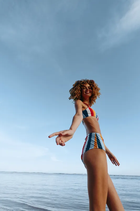 Confident woman in a colorful swimsuit stands by the ocean at the beach during summer under a clear blue sky curly hair blowing relaxed freedom mood soft natural light bright sunlight