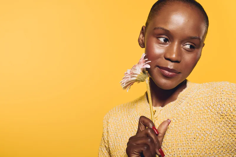 Young black woman with dark skin holding a pink flower looks to the side against a yellow background. Concept of natural beauty, skincare, and serene mood in studio lighting.