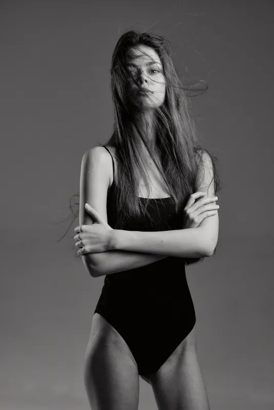 Black and white portrait of a young woman in a black bodysuit with long hair, posing with crossed arms and a calm expression.