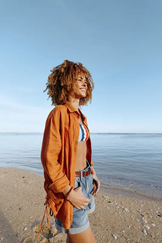 A young brunette woman on a sunny sandy beach stands in a relaxed pose, wearing an orange shirt over a striped bikini top and denim shorts, smiling as a light sea breeze blows through her hair.