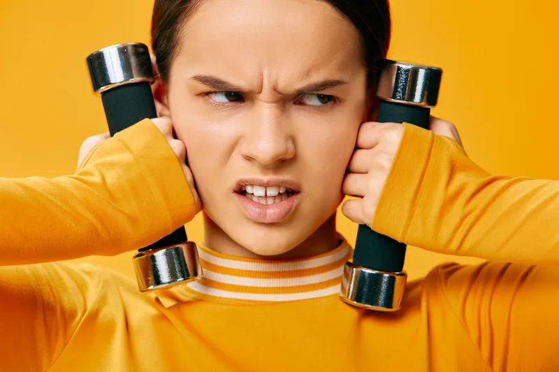 Fitness woman holds dumbbells near her ears in a bright orange studio, delivering an intense workout. A focused athlete shows strength, balance and determination during training.