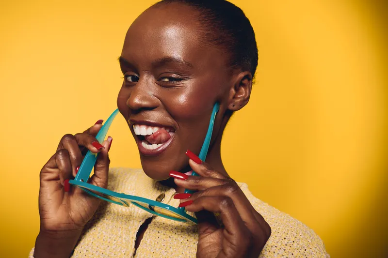 Smiling dark skinned African woman with short hair holds turquoise sunglasses and smiles playfully. Yellow background creates bright cheerful mood. Concept of beauty, fashion, and confidence.