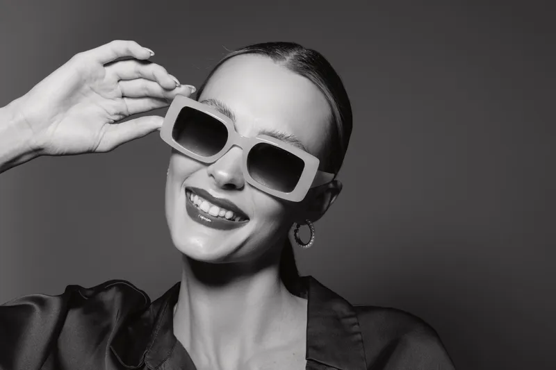 Smiling Caucasian woman with pale skin wearing large sunglasses and hoop earrings poses confidently on dark background. Concept of fashion, style, and happiness.