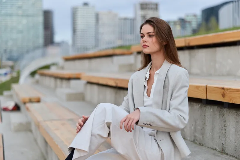 business woman in light gray suit sitting on urban stairs with pensive expression, professional outfit, city background, confident female in modern style