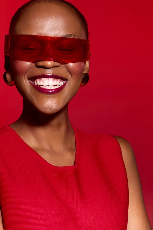 Smiling African woman with dark skin wearing red sleeveless top and red translucent sunglasses on red background. Concept of fashion, beauty, confidence, and vibrant style.