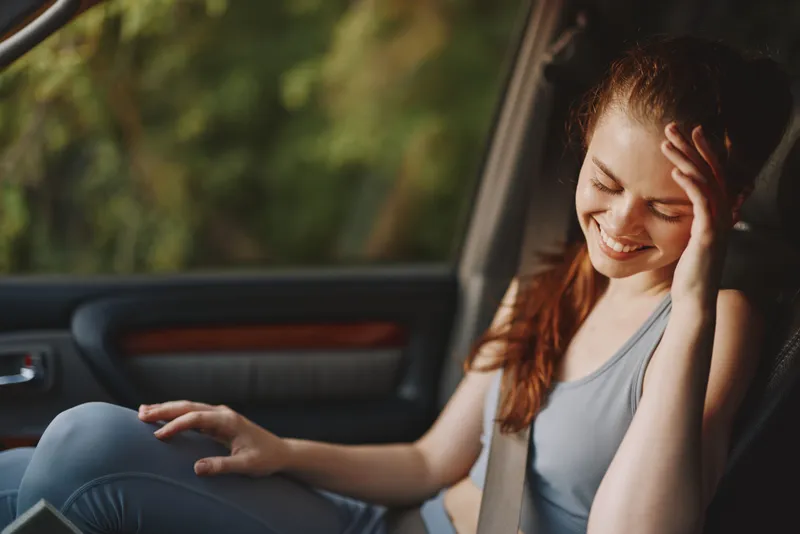 Young woman enjoying a moment of relaxation in a car, wearing sporty clothes, smiling and looking cheerful, surrounded by nature in a serene outdoor setting