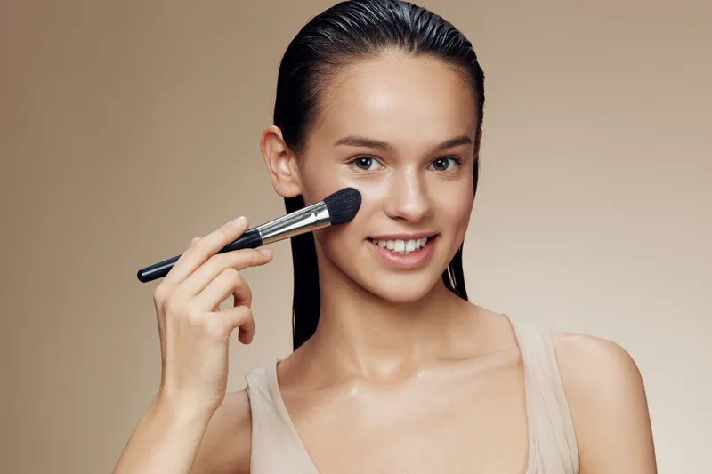 Young woman applying makeup with a brush, showcasing a radiant smile and natural beauty, perfect for beauty and skincare concepts