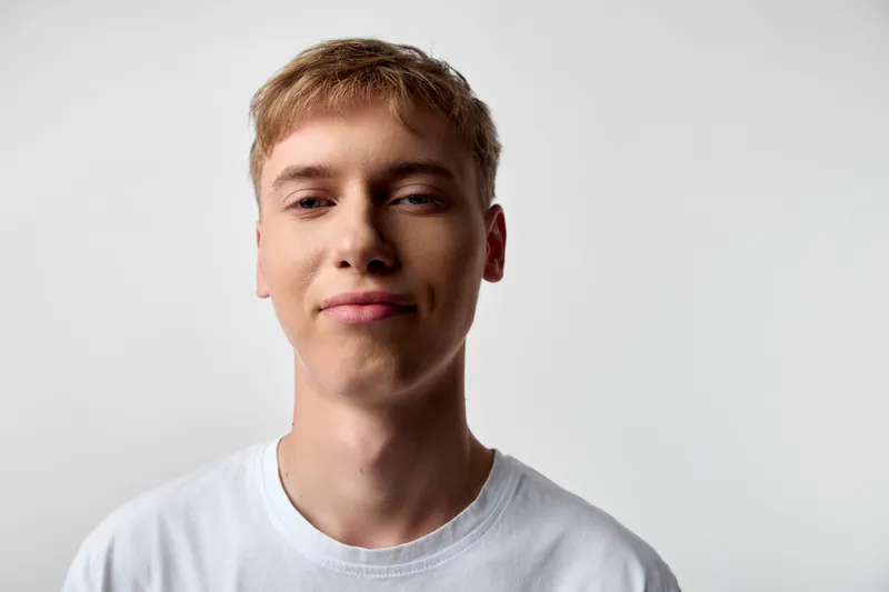 Young man with short blond hair and a subtle smile wearing a white tshirt, posing against a neutral background, conveying confidence, simplicity, and a friendly, approachable vibe.