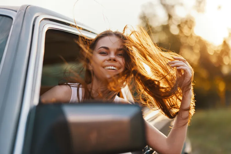 Cheerful woman enjoying a summer road trip with hair blowing in the wind, capturing the essence of freedom and adventure in a sunny outdoor setting.