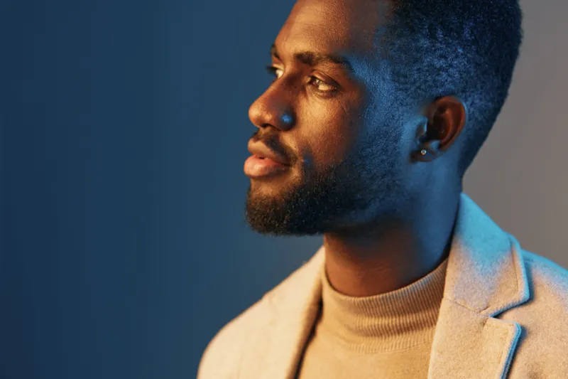 Portrait of a stylish young Black man in a beige suit jacket, showcasing confidence and sophistication against a blue and orange gradient background