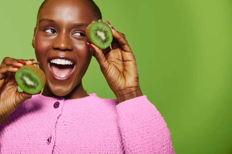 Happy African woman with dark skin holds two halves of fresh kiwi fruit near face on green background. Concept of healthy eating, natural food, and vibrant lifestyle with bright pink sweater.