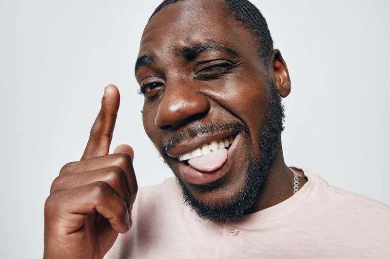 African American man with dark skin smiling and winking while pointing finger up in casual pink shirt on white background. Positive expression and playful gesture closeup.