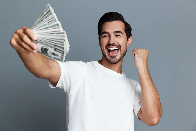 Happy smiling man holding fan of cash celebrating his success with joyful expression, wearing casual white t-shirt against a neutral grey backdrop.