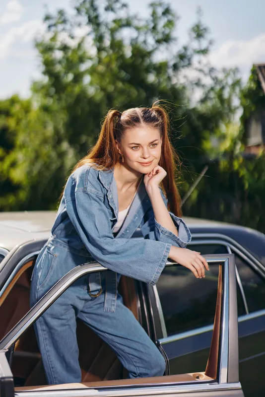 Young woman in denim outfit leaning on car, showcasing a casual and joyful mood outdoors in a vibrant summer setting with greenery.
