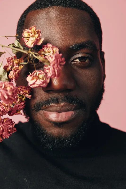 African man with dark skin and beard posing with dried pink flowers near face in creative conceptual visual style on pink background. Portrait with artistic expression and floral detail.
