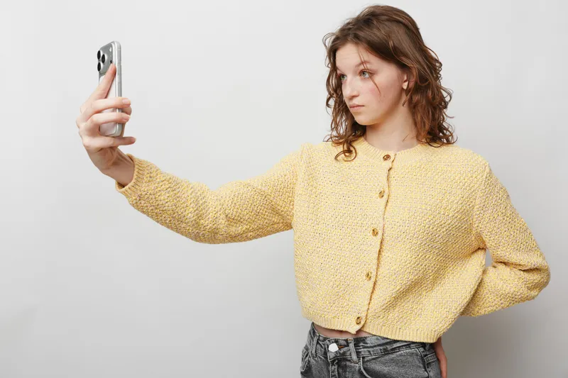Young woman with curly hair wearing yellow knitted sweater takes selfie with smartphone on white background. Casual style, social media concept, modern technology and communication.