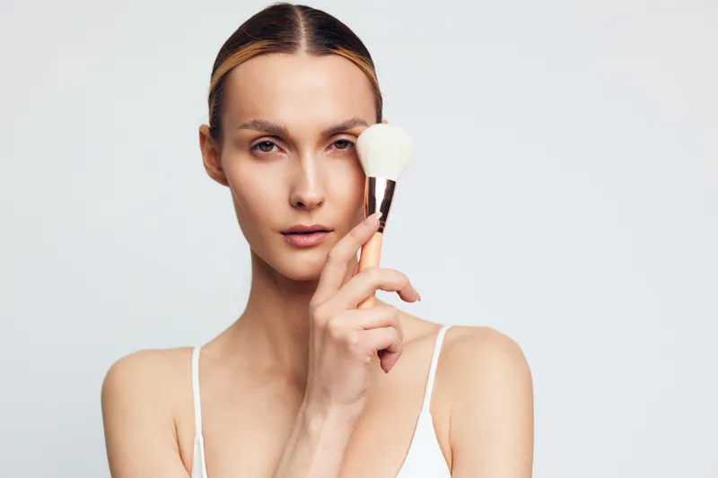 Young woman with clear skin holding makeup brush near face on white background. Concept of beauty, skincare, and natural makeup routine. Soft lighting highlights facial features and smooth complexion.
