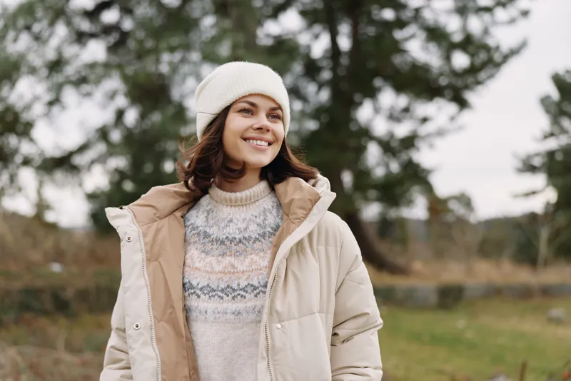 Woman in a warm coat and knit sweater outdoors, smiling under a soft beanie in a chilly landscape, casual autumn fashion and outdoor style featuring cozy layers
