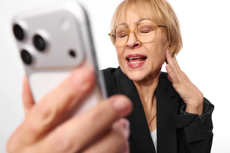Elderly woman with glasses reacts during a smartphone call, hand at ear, showing hearing difficulty, stress and frustration. Blurred mobile device suggests remote communication.