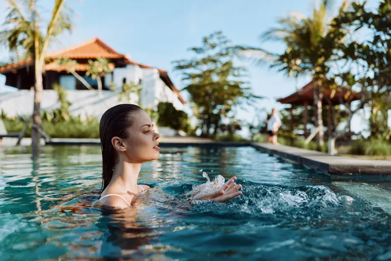 Relaxed woman in a pool enjoys a sunlit moment with gentle ripples, tropical resort backdrop. Lifestyle scene highlighting wellness, leisure, and self care in a warm outdoor setting.