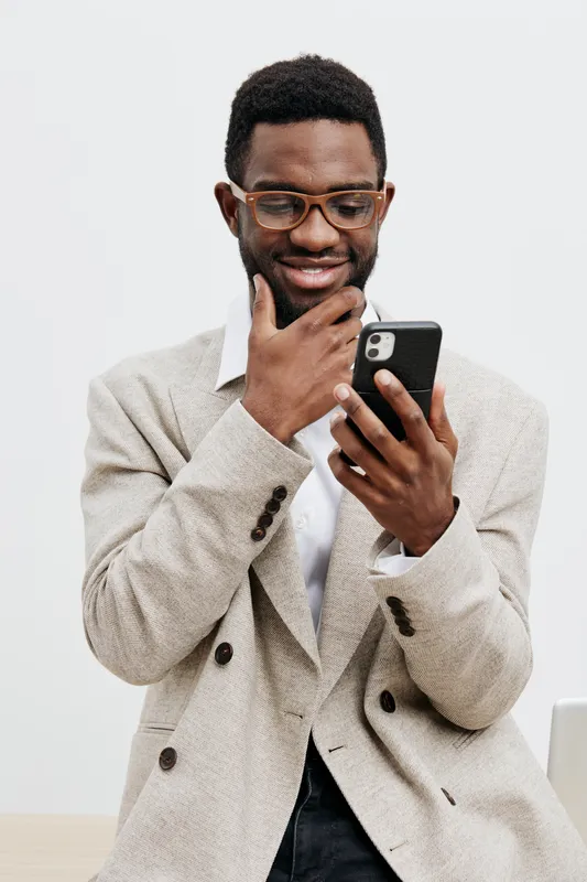 Smiling man using smartphone, casually dressed in beige blazer and glasses, looking happy and engaged with device, indoor studio shot, positive mood, modern lifestyle, technology concept, diversity.