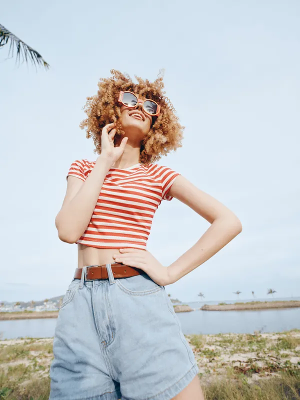 Young woman with curly hair wearing a striped crop top and high waisted denim shorts stands outdoors under a bright sky, posing confidently with sunglasses and a hand on hip.