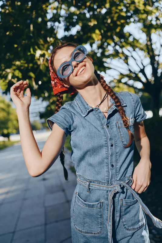 Lifestyle woman in modern boho-western denim street style wears large sunglasses and red bandana, smiling outdoors on sunny day with vibrant film color tones.