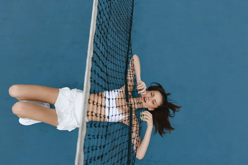 Young athletic woman lying on a tennis court, showcasing sporty style in white outfit, conveying joy and relaxation under a bright blue sky