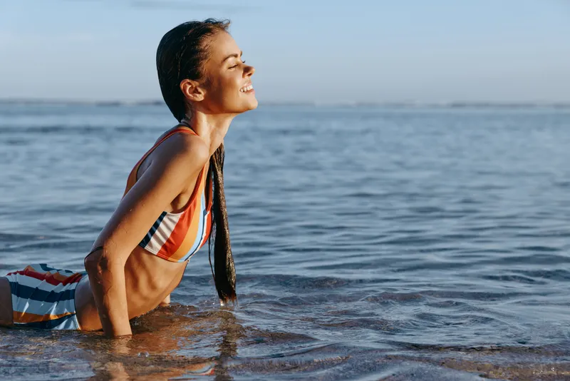 Young woman in striped swimsuit enjoys sunny day at the beach, lying in shallow water with eyes closed and smiling, feeling relaxed and peaceful by the sea.