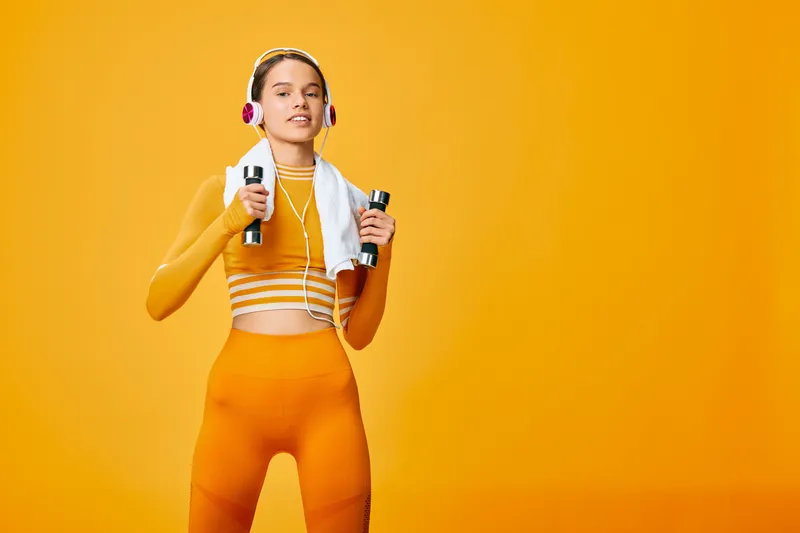 Fitness woman in bright orange sportswear stands in a studio, holding water bottles with a towel over her shoulders and wireless headphones, ready for a workout session.