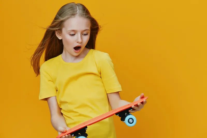 Young girl in a yellow shirt surprised by her skateboard against a vibrant yellow background, conveying joy and excitement in a playful, energetic setting