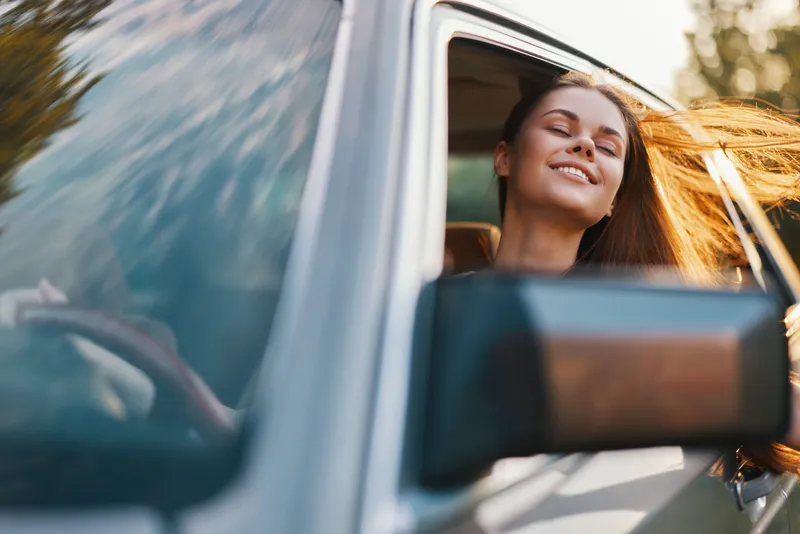 Young woman enjoying a sunny drive with her hair flowing in the wind, expressing happiness and freedom in a car, perfect for concepts of adventure and joy.
