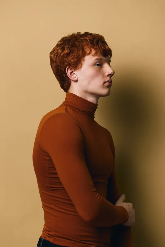 Young man with red hair wearing brown turtleneck standing sideways against beige background. Profile portrait of thoughtful male model with fair skin and casual style indoors.