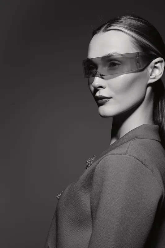 Portrait of a young white woman with light skin wearing futuristic glasses and textured jacket in black and white studio lighting.