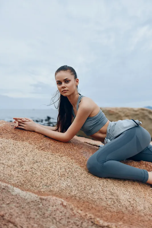 Young athletic woman in sportswear posing on rocky beach, showcasing strength and focus with a calm sea backdrop, emphasizing fitness and health lifestyle