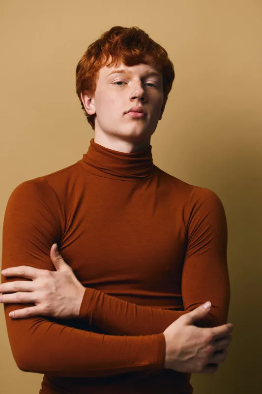 Young man with red hair wearing brown turtleneck sweater posing with crossed arms against beige background. Confident male model with fair skin and casual style portrait.