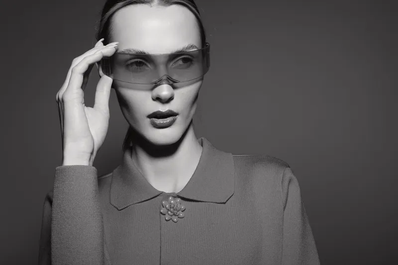 A young white woman with pale skin adjusts oversized translucent sunglasses, wearing a textured sweater with a collar and brooch. Studio portrait with dramatic lighting and neutral background.