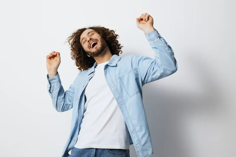 Happy young man with curly hair dancing joyfully in a light blue shirt, expressing excitement and positivity against a minimalistic white background