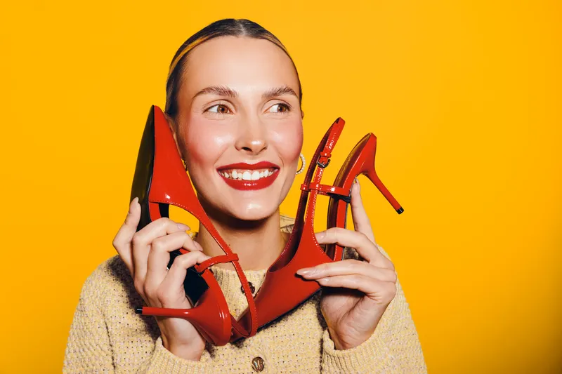 Smiling woman holds red high heel shoes near face on bright yellow background. Concept of fashion, style, footwear, and happiness with vibrant colors and joyful expression.