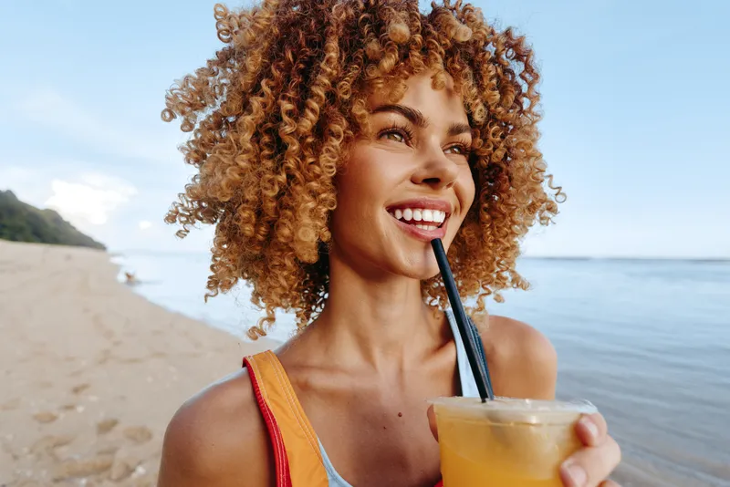 Smiling woman with curly hair on the beach in summer holding an iced juice drink with a straw while enjoying outdoors by the seaside shoreline natural light summer vacation mood relaxed