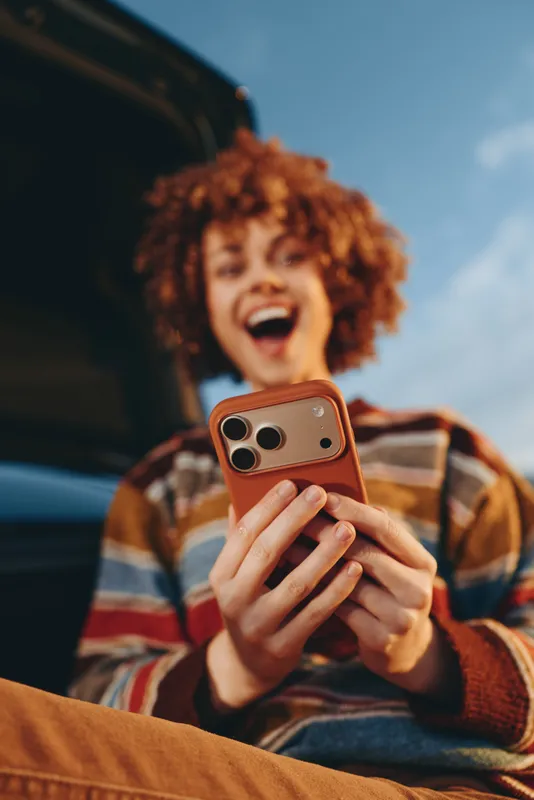 Curly haired child smiling while holding smartphone inside a car, lifestyle travel moment, outdoor sky background, casual sweater, candid portrait, young cheerful expression