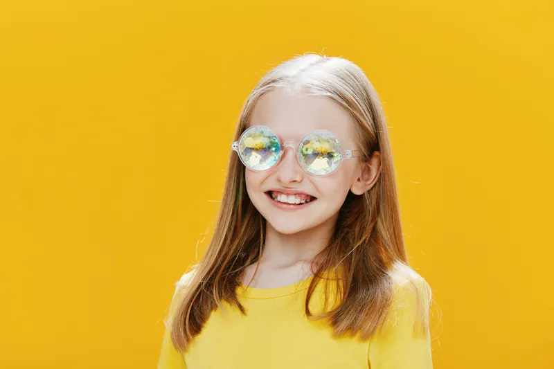Cheerful young girl wearing colorful glasses and a yellow shirt, smiling against a vibrant yellow background, showcasing happiness and playfulness