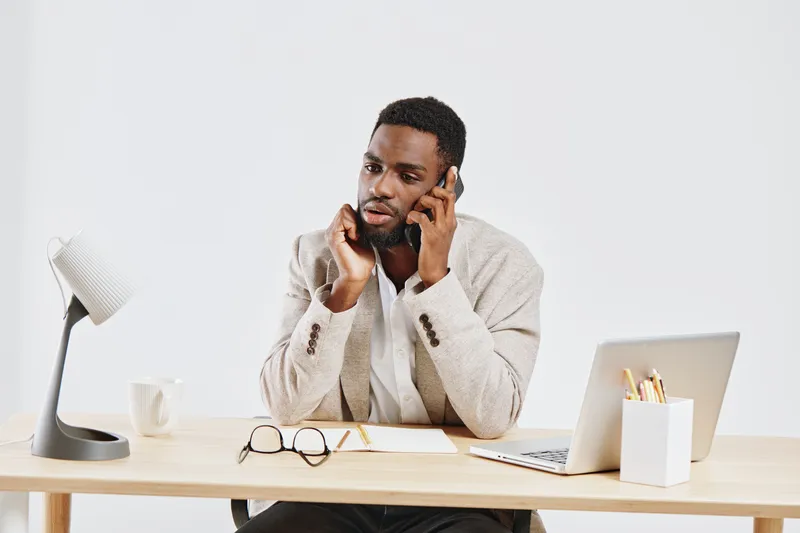 Professional man talking on mobile phone while working at minimalist office desk with laptop, glasses, and stationery; workspace with clean, bright background; business communication concept.
