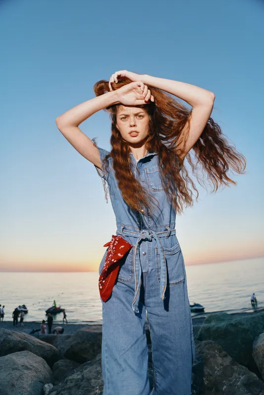 Lifestyle woman in modern boho-western denim street style stands by seaside at sunset with film color tones capturing natural casual fashion mood and texture.