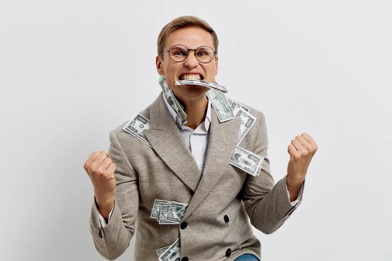 Frustrated angry man in business suit with glasses, holding crumpled dollar bills in his mouth, looking upset, isolated on plain white background, studio portrait, financial stress concept.